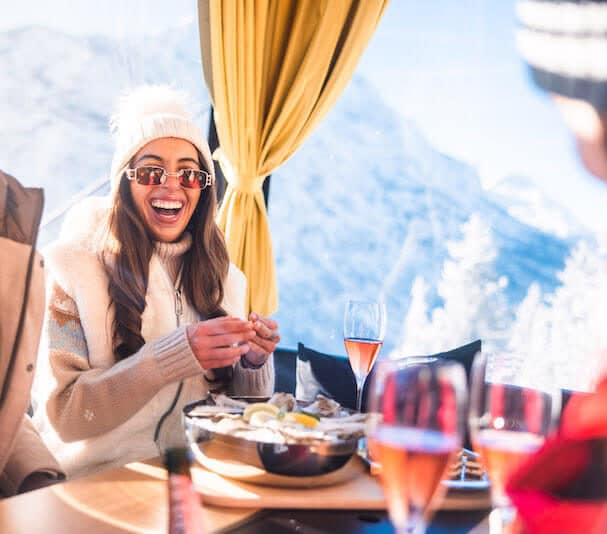 Eine Skifahrerin sitzt lachend in der Moet Chandon Champagner Gondel. Vor ihr ist der Tisch mit diversen Speisen gedeckt.