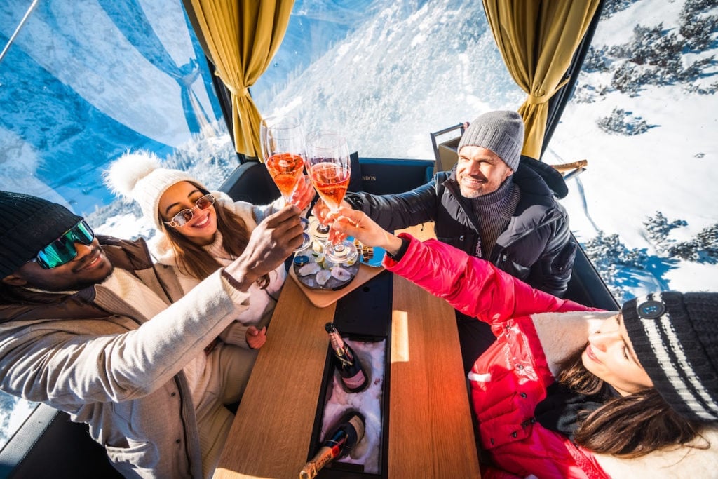 Zwei Skifahrerinnen und zwei Skifahrer sitzen in der Gondel und stoßen lachend Gläser zum Prosten zusammen.