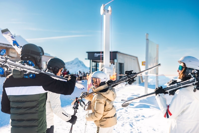 Eine kleine Gruppe Skifahrerinnen und Skifahrer gehen zur Gondelstation und lachen dabei in die Kamera.